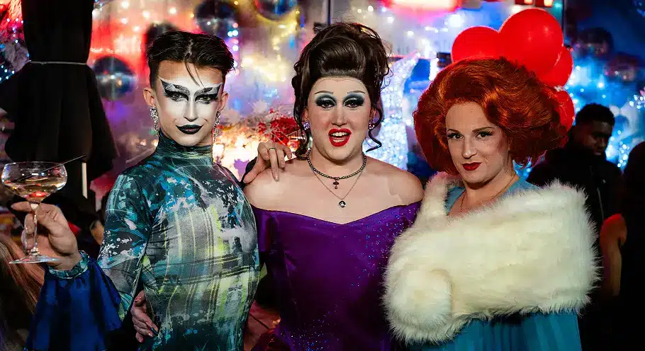 Three drag hosts posing together at Arch Clapham during a live drag night, surrounded by colourful lights and party decorations at the popular gay bar in London.
